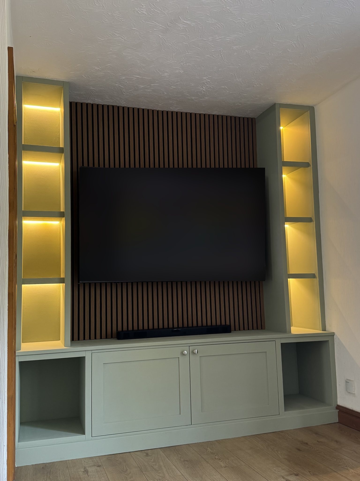 A sage green media wall with LED-lit shelves either side, slatted oak acoustic panelling and shaker cupboards below