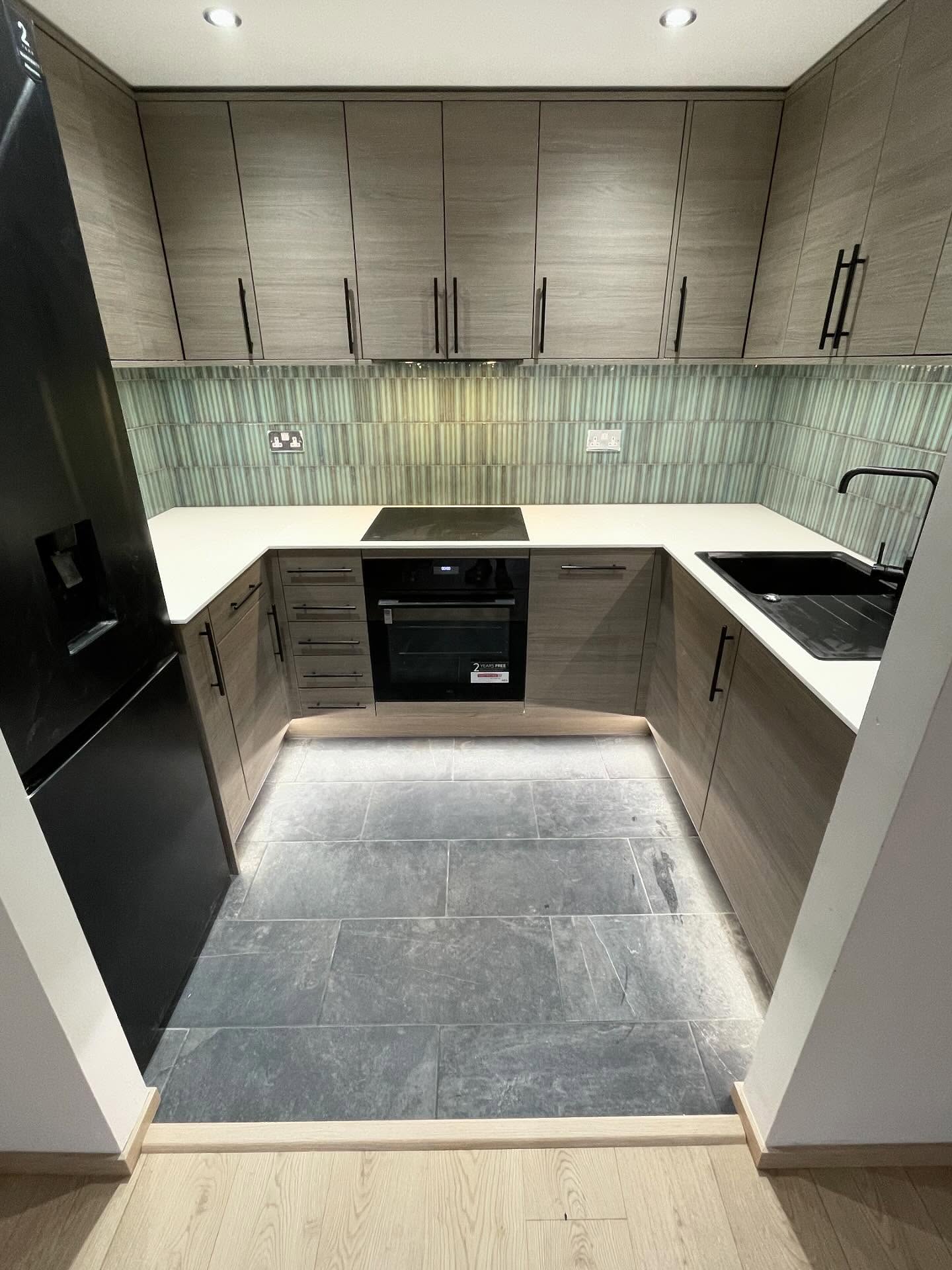 A handleless woodgrain kitchen with a white worktop, green tiled splashback and slate floor