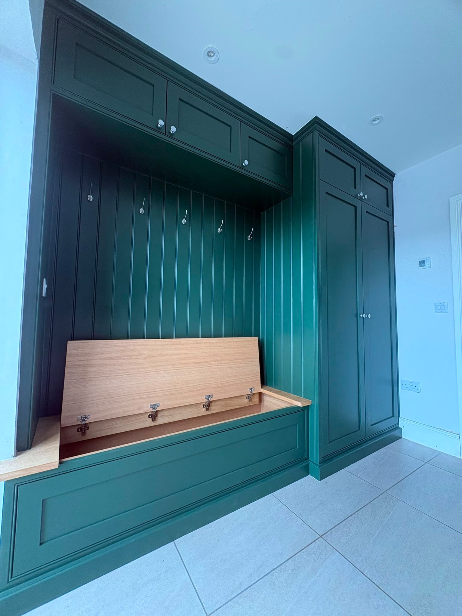 A green boot room with tongue and groove panelling, coat hooks, tall cupboards and an oak lift-up bench