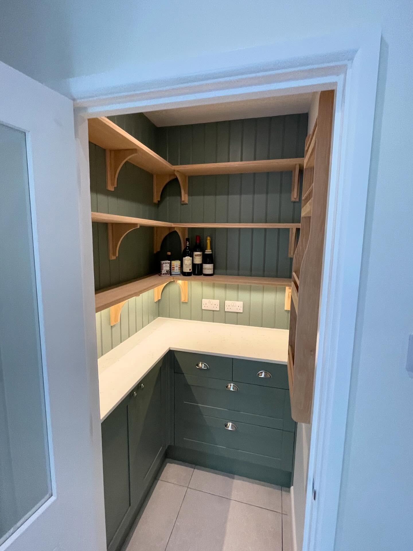 A walk-in pantry with green shaker units, oak shelves on curved brackets, wine racks and under-cabinet lighting