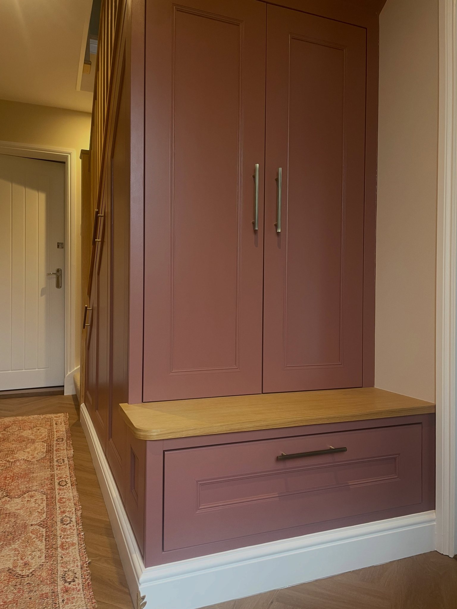 Under-stairs storage painted in a muted pink, with an oak bench top and a pull-out shoe drawer